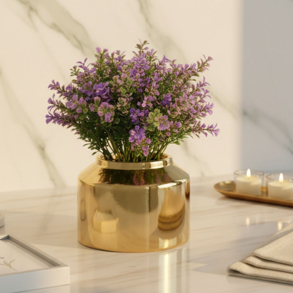 A shiny gold cylindrical vase filled with small purple flowers and green leaves, placed on a white marble surface against a white marble wall. Two lit candles in small glass holders sit on a tray in the background.