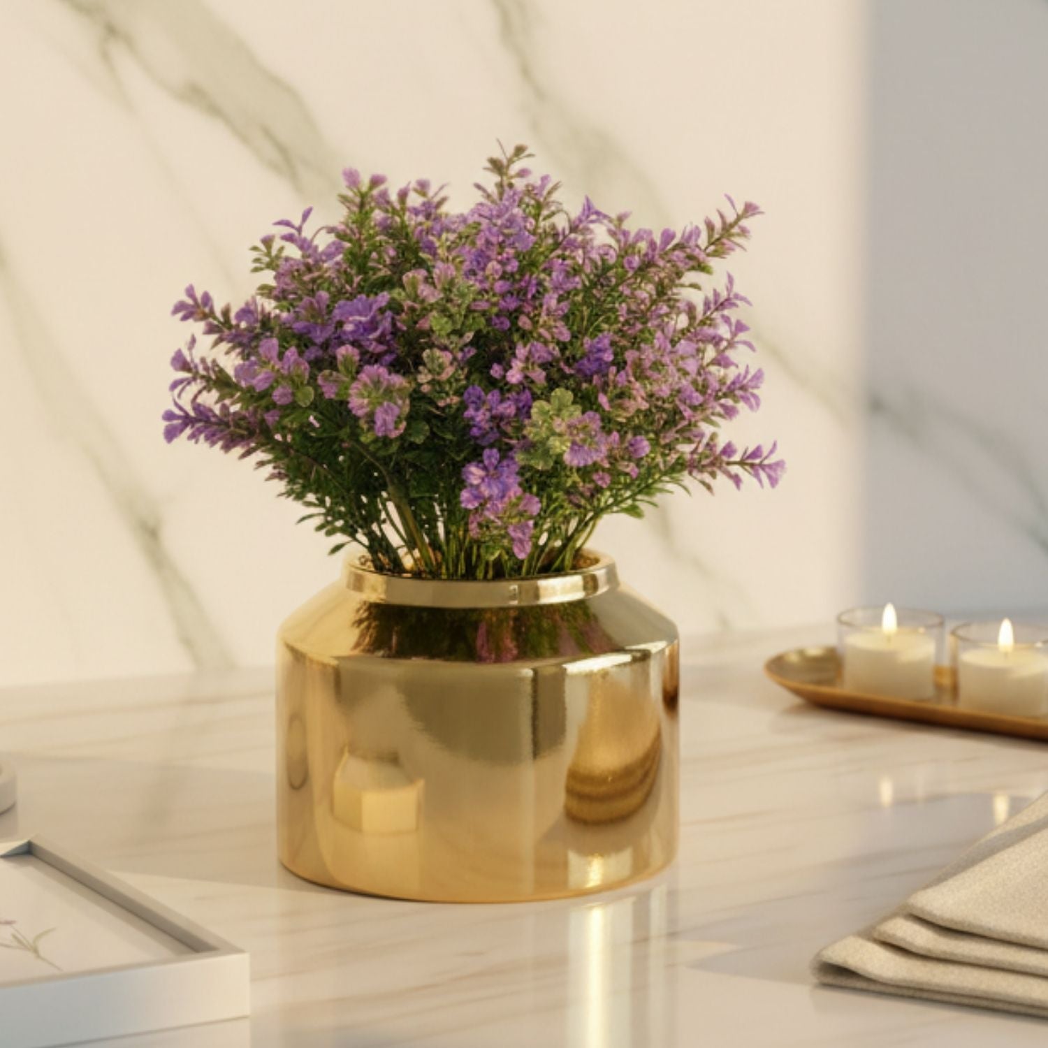 A shiny gold cylindrical vase filled with small purple flowers and green leaves, placed on a white marble surface against a white marble wall. Two lit candles in small glass holders sit on a tray in the background.