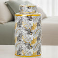 A cylindrical ceramic jar with a matching lid, decorated with a pattern of black and gold tropical leaves on a white background, set on a light-colored table in front of a blurred sofa.