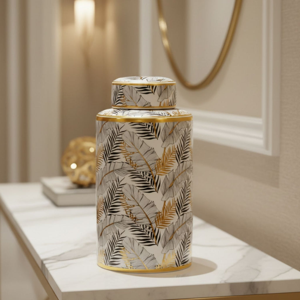A decorative cylindrical jar with a lid, featuring a white base with a black and gray palm leaf pattern and metallic gold accents, displayed on a marble console table in a warm, elegant interior setting.