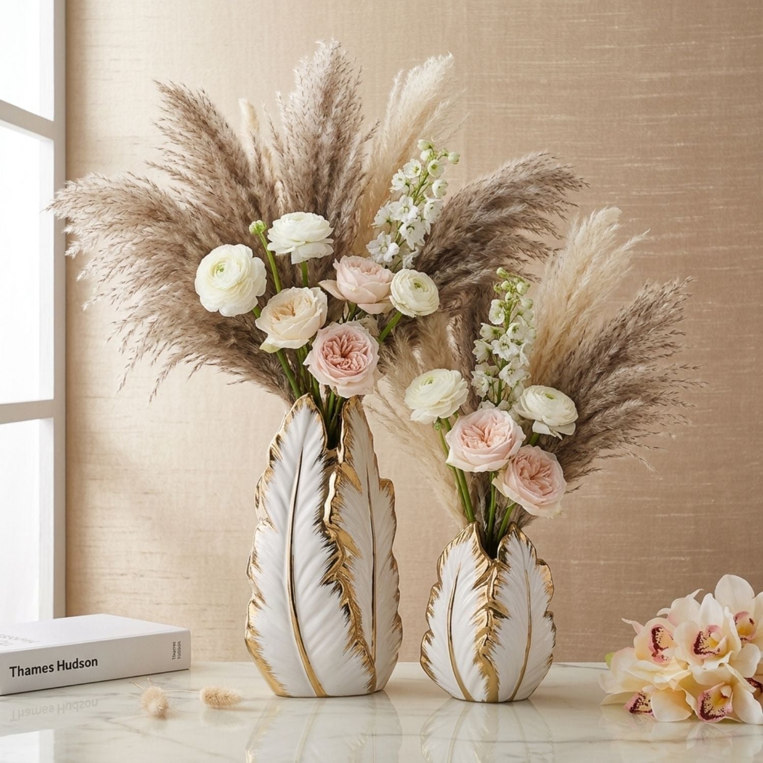 Two decorative leaf-shaped ceramic vases with gold veining, filled with pampas grass and blush pink and white flowers, displayed on a tabletop against a beige wall.