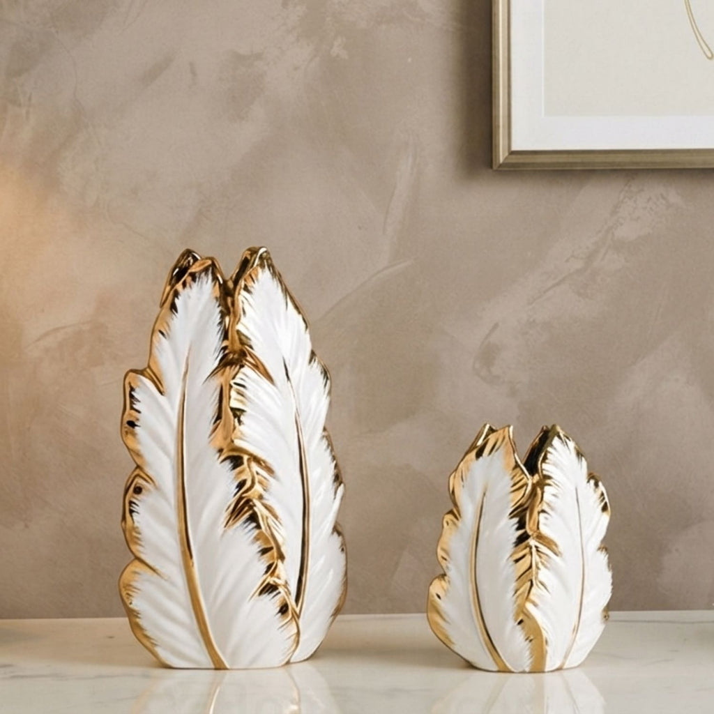 Two decorative feather-shaped vases in white with metallic gold trim, displayed on a light marble surface against a textured beige wall.