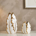 Two decorative feather-shaped vases in white with metallic gold trim, displayed on a light marble surface against a textured beige wall.