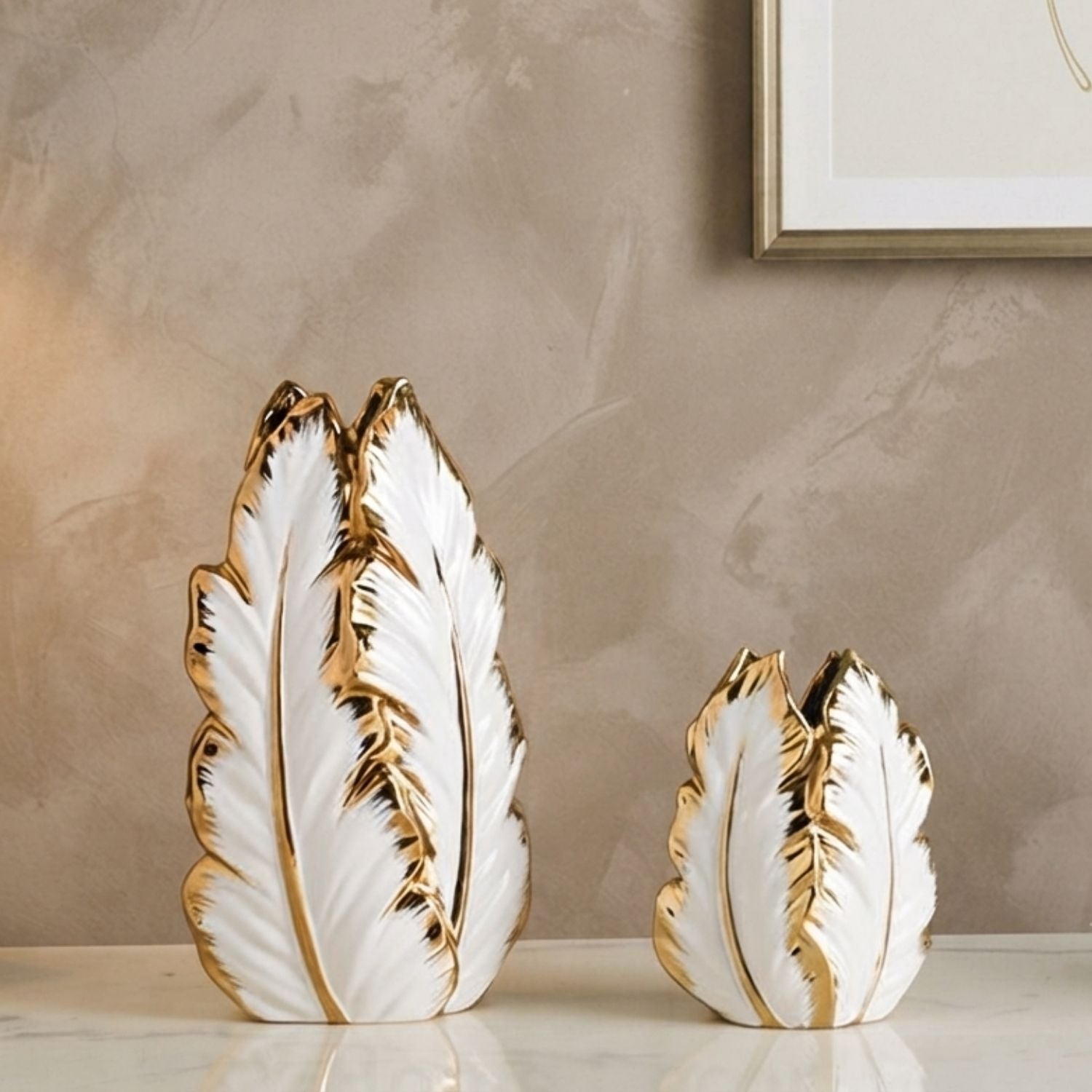 Two decorative feather-shaped vases in white with metallic gold trim, displayed on a light marble surface against a textured beige wall.