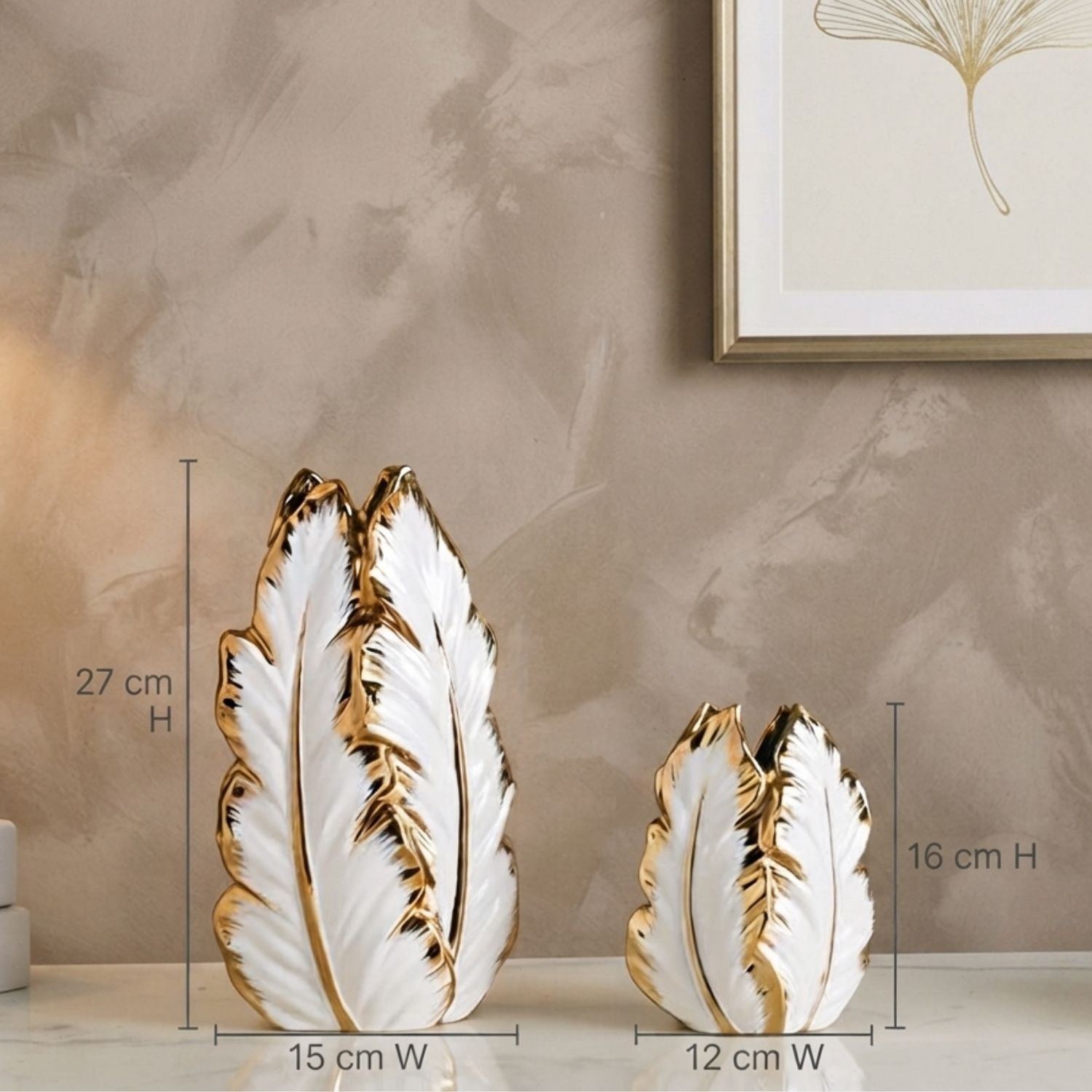 Two decorative leaf-shaped ceramic vases in white with gold trim, shown on a tabletop against a textured beige wall. Measurement lines and text indicate the larger vase is 27 cm high by 15 cm wide, and the smaller vase is 16 cm high by 12 cm wide.