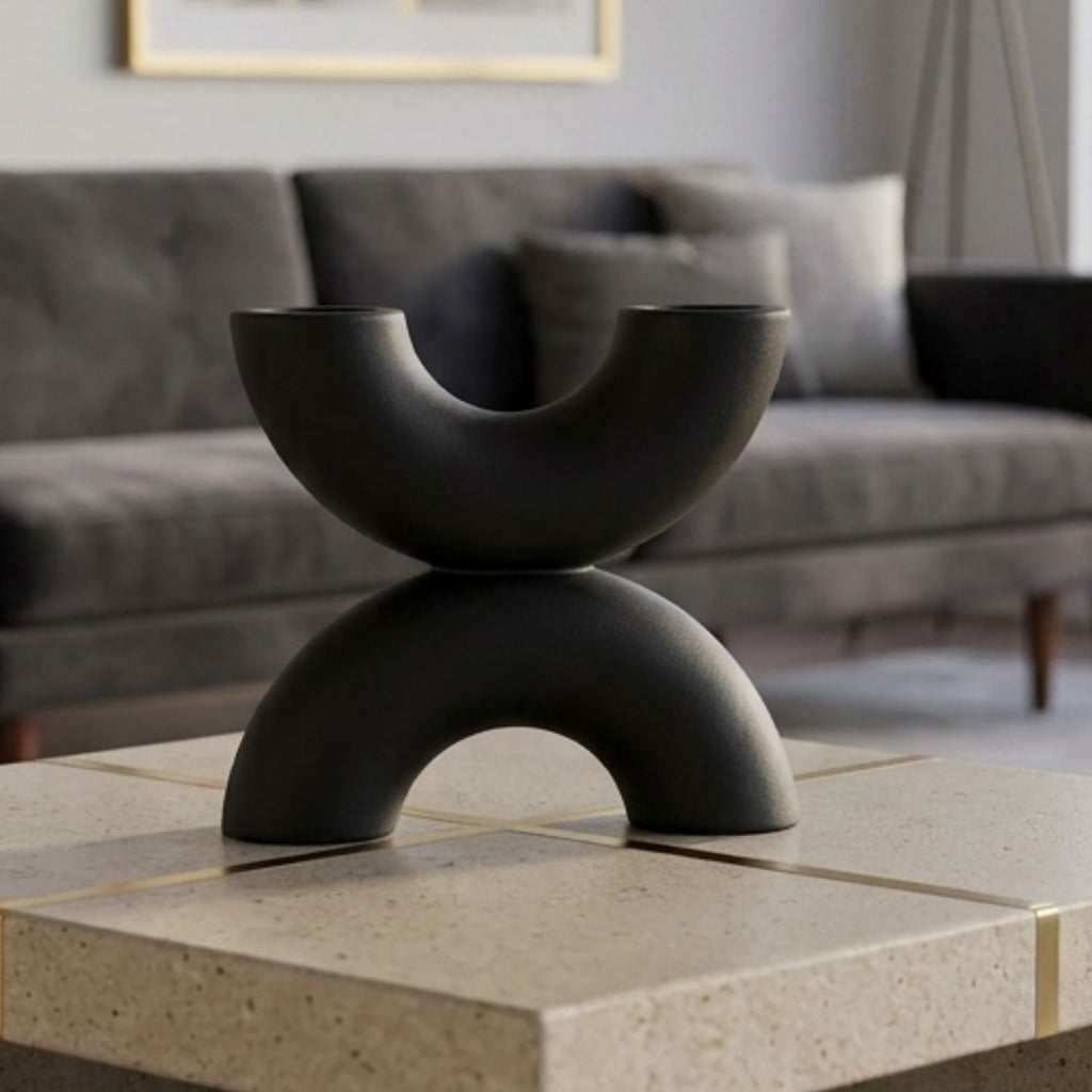 A matte black sculptural two-tiered bowl stand displayed on a light stone tile table in a modern living room setting with a gray sofa in the background.