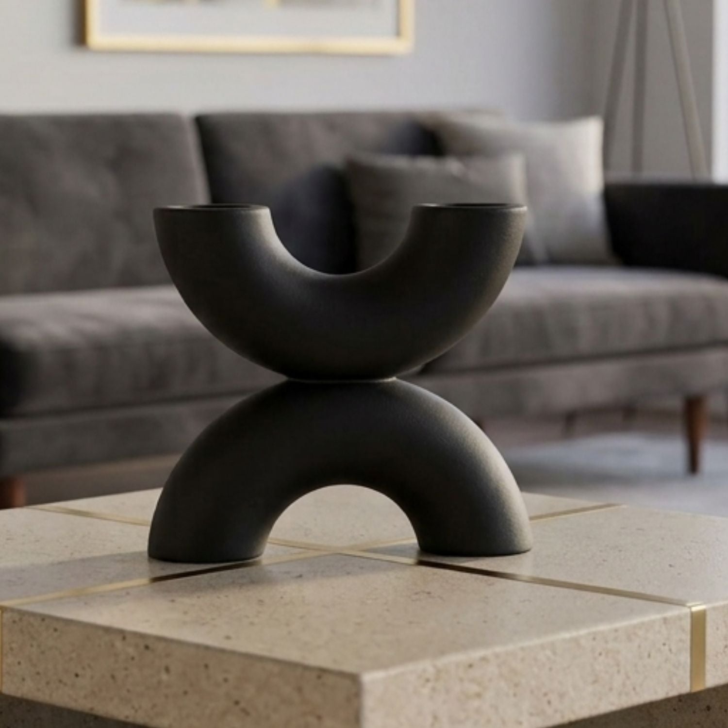A matte black sculptural two-tiered bowl stand displayed on a light stone tile table in a modern living room setting with a gray sofa in the background.