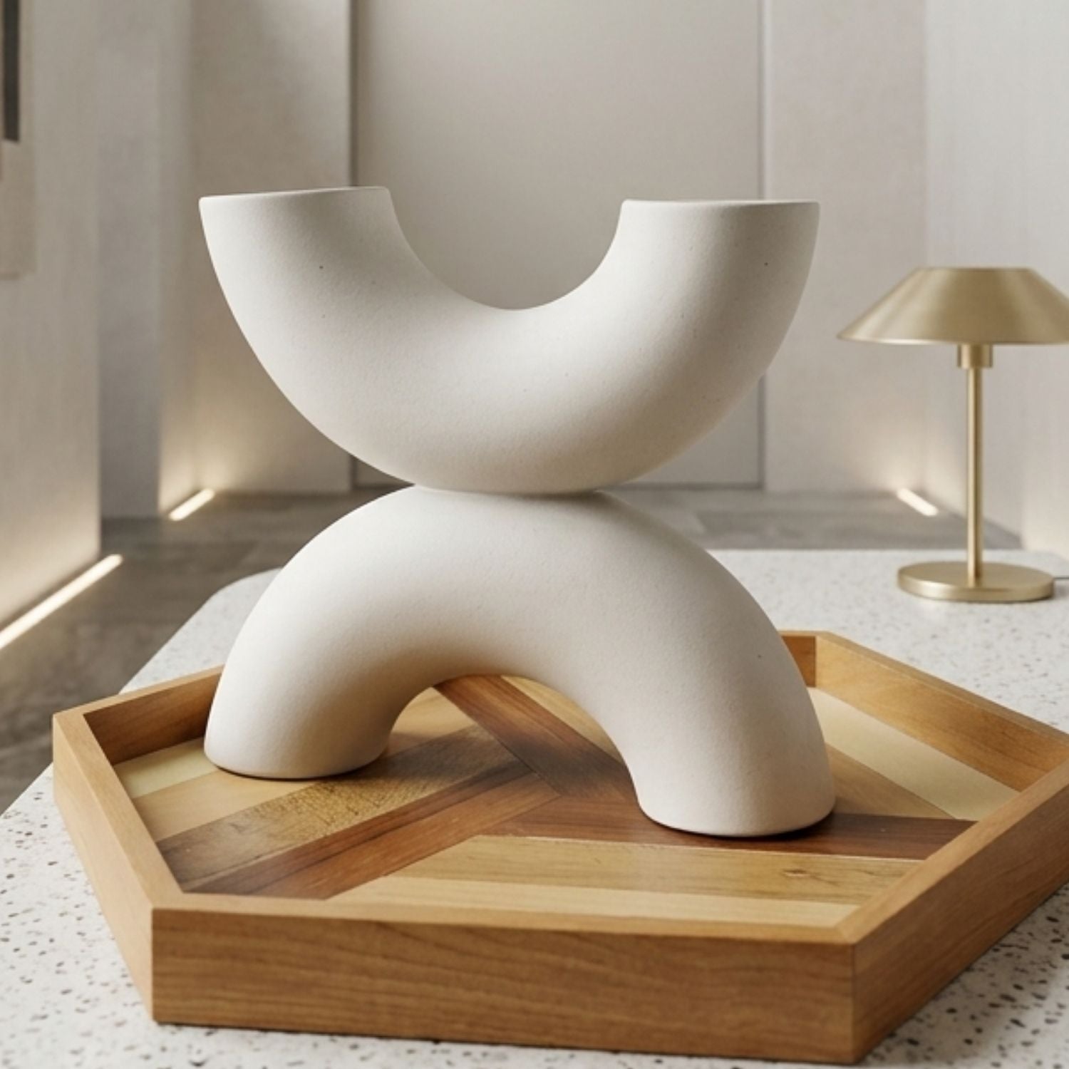 A modern abstract ceramic vase in a matte off-white finish, styled on a hexagonal wooden tray with a gold lamp in the background.