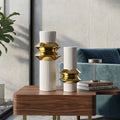 Two modern decorative vases with white ribbed cylindrical bodies and layered metallic gold geometric fluted tops, displayed on a wooden side table in a styled interior setting with a blue sofa, books, and a plant.