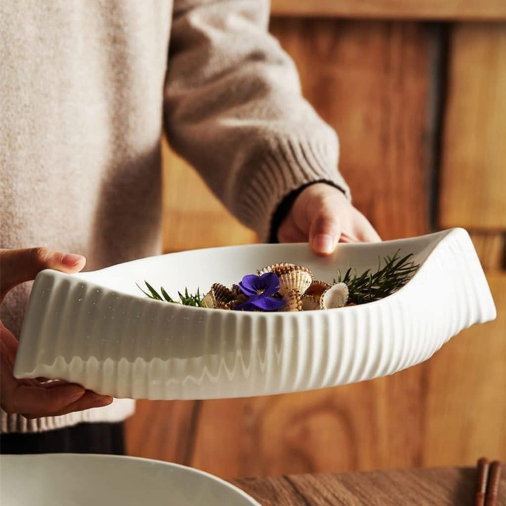 A person in a beige sweater holds a white ribbed ceramic bowl or dish containing small seashells, greenery, and a purple flower, against a warm wooden background.