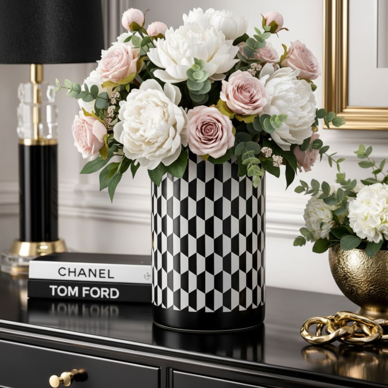 A black and white geometric patterned cylindrical vase filled with white and blush pink artificial flowers and green foliage, displayed on a glossy black console table next to a black lamp and decorative books.