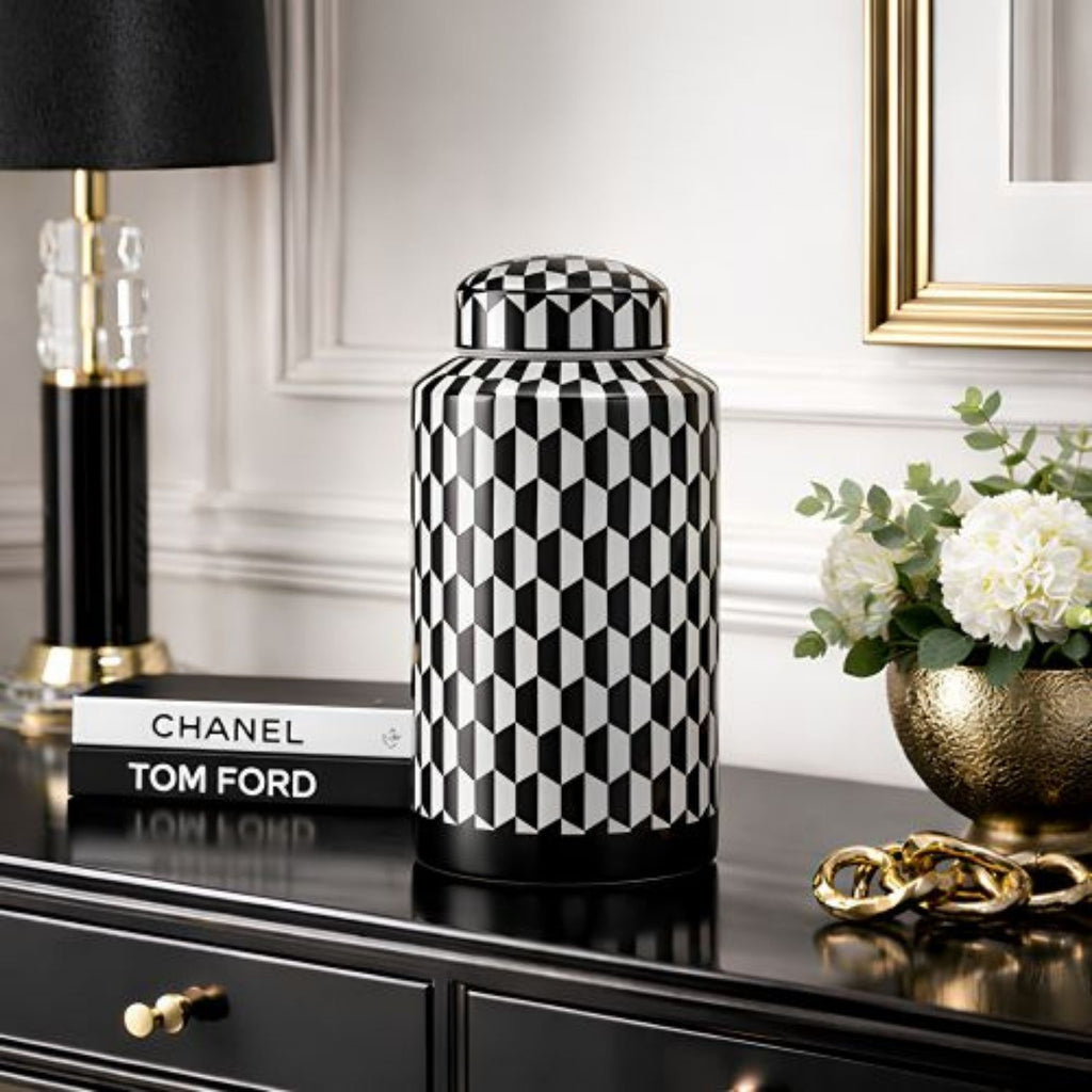 A black and white geometric patterned ceramic jar with a matching lid, styled on a black console table next to a black and gold table lamp, a Chanel and Tom Ford book, and a gold vase of white flowers.