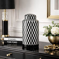 A black and white geometric patterned ceramic jar with a matching lid, styled on a black console table next to a black and gold table lamp, a Chanel and Tom Ford book, and a gold vase of white flowers.
