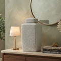 A large textured light gray ceramic jar with a matching lid sits on a wooden console table in a styled interior setting, next to a lit table lamp and books, with a round mirror on the wall behind it.