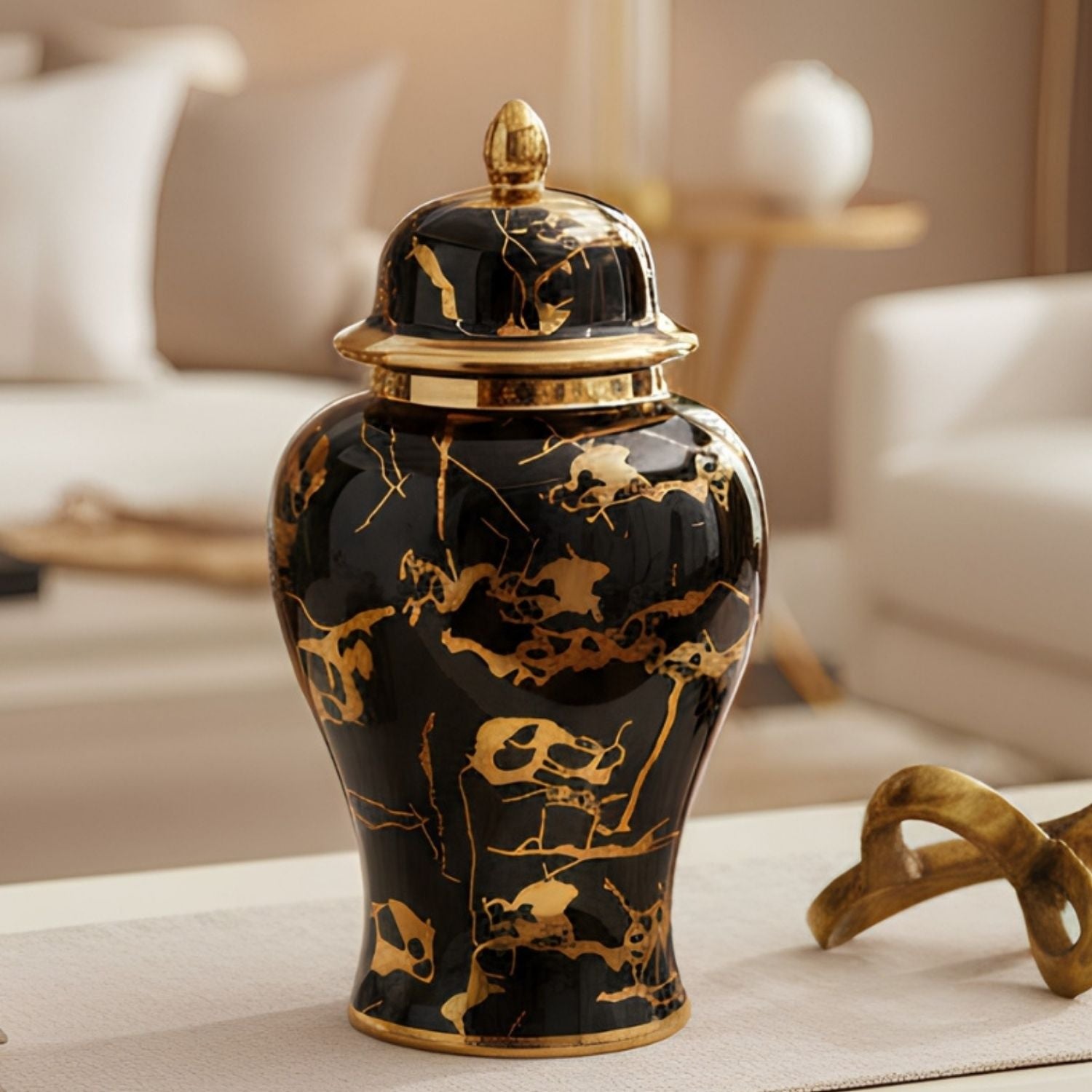 A glossy black ceramic jar with a matching lid, decorated with intricate gold marble-like veining and gold trim, set on a light beige table in a softly blurred living room background.