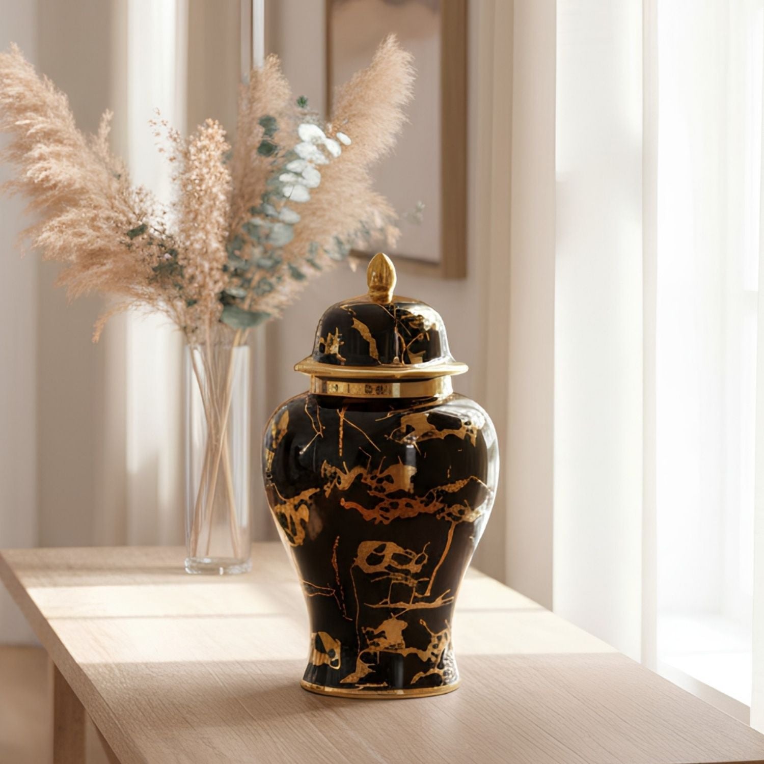 A black ceramic decorative jar with gold marble-style veining and a matching lid, displayed on a light wood table in a softly lit interior setting with dried grasses in a vase behind it.