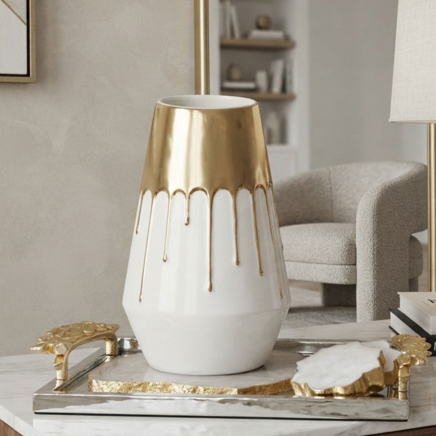 A white ceramic vase with a metallic gold drip design on the upper half, displayed on a marble tray in a modern living room setting.