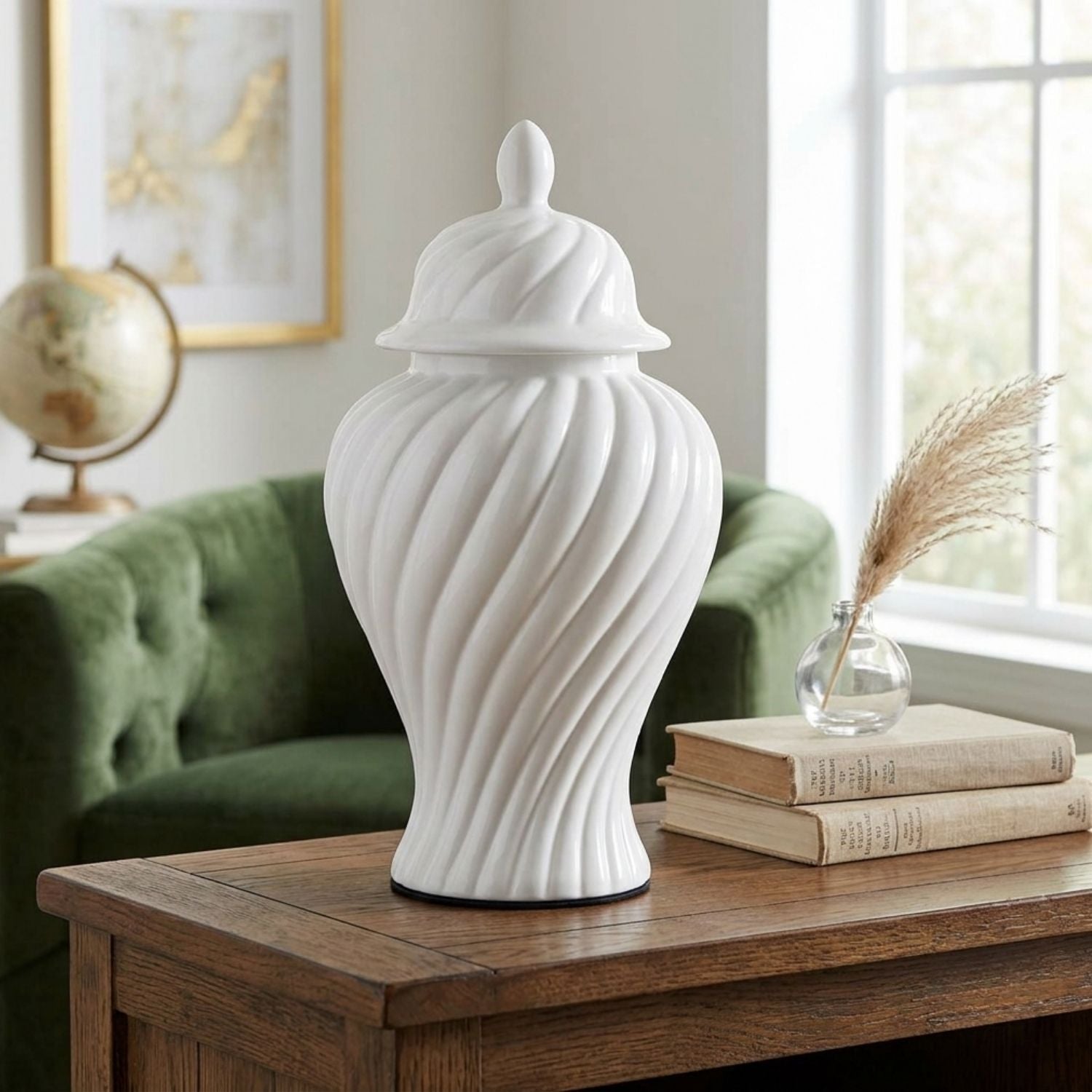 A white ceramic lidded jar vase with a fluted, ribbed texture stands on a wooden table in a styled living room setting next to books and a small glass vase with dried grass.
