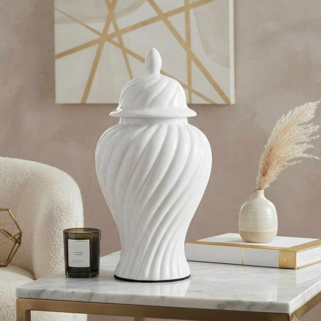 A white ceramic lidded jar with a ribbed spiral texture, displayed on a marble-topped side table in a styled room setting next to a candle, books, and a small vase.