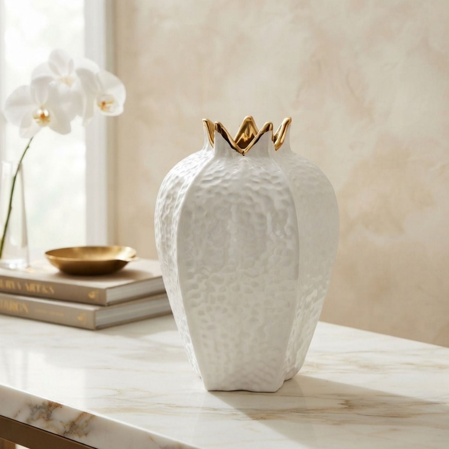 A white textured ceramic vase with a gold-leaf crown rim sits on a white marble console table. In the background, a stack of books and a vase of white orchids are visible against a warm beige wall.