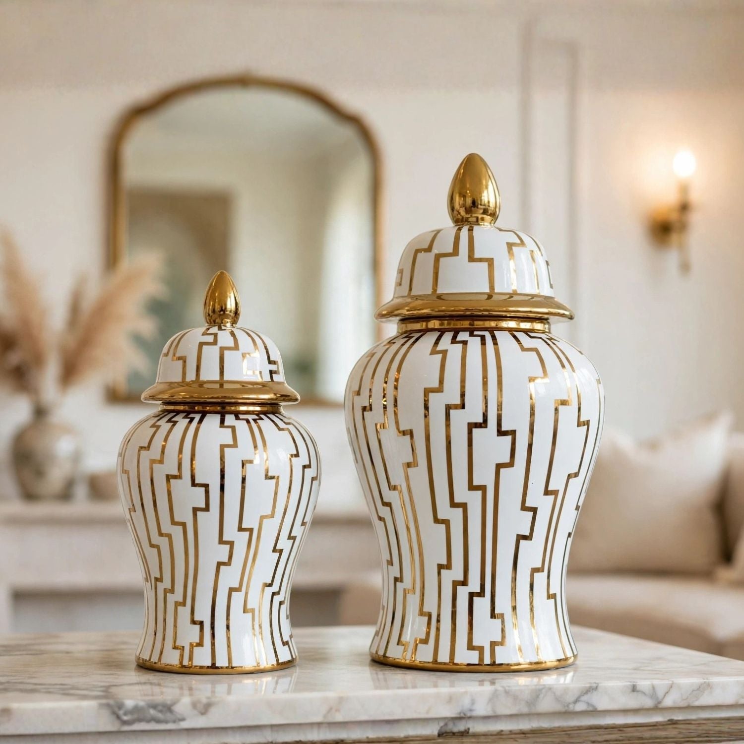 Two white ceramic decorative canisters with gold geometric line patterns and gold finials, displayed on a marble surface in a softly lit interior setting.