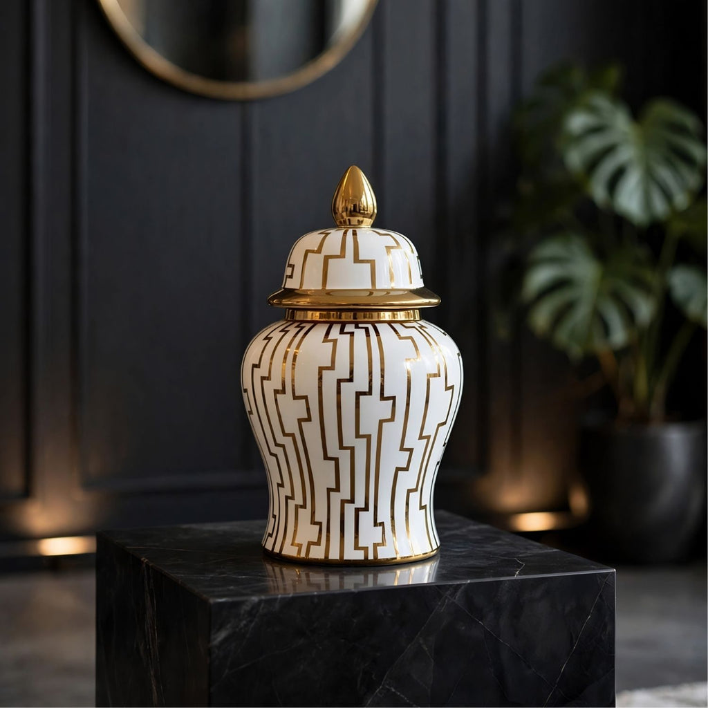 A decorative ceramic ginger jar with a white body, gold geometric pattern, and gold lid finial, displayed on a black marble pedestal in a dark interior setting.
