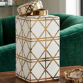 A white ceramic decorative canister with a geometric gold line pattern and a matching lid, displayed on a wooden table in front of a green velvet armchair.