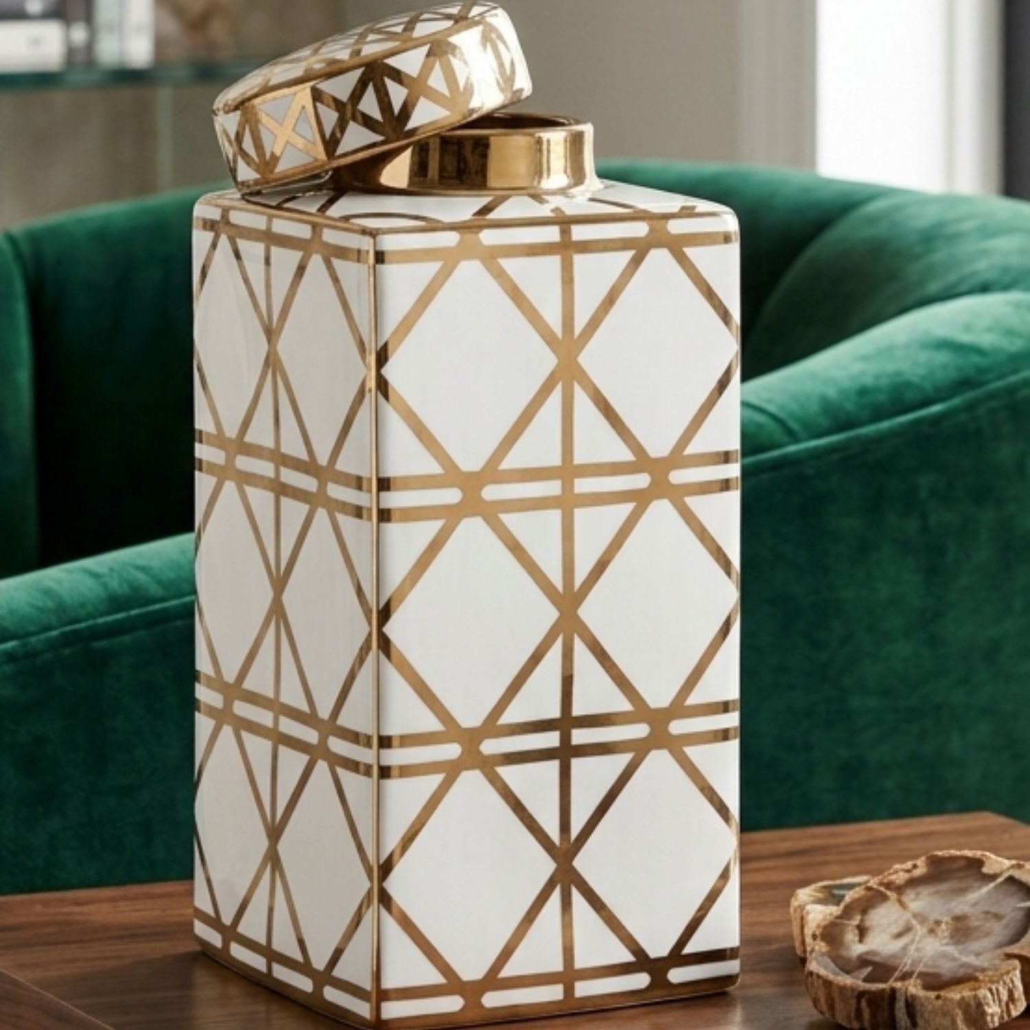 A white ceramic decorative canister with a geometric gold line pattern and a matching lid, displayed on a wooden table in front of a green velvet armchair.