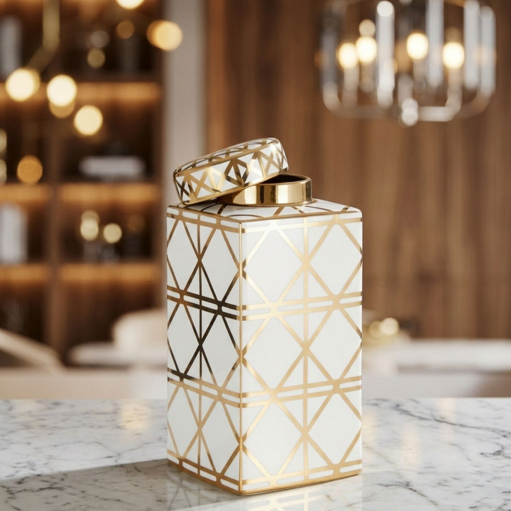 A white ceramic canister with a gold geometric line pattern and a matching lid, displayed on a marble countertop in a warm, blurred interior setting.