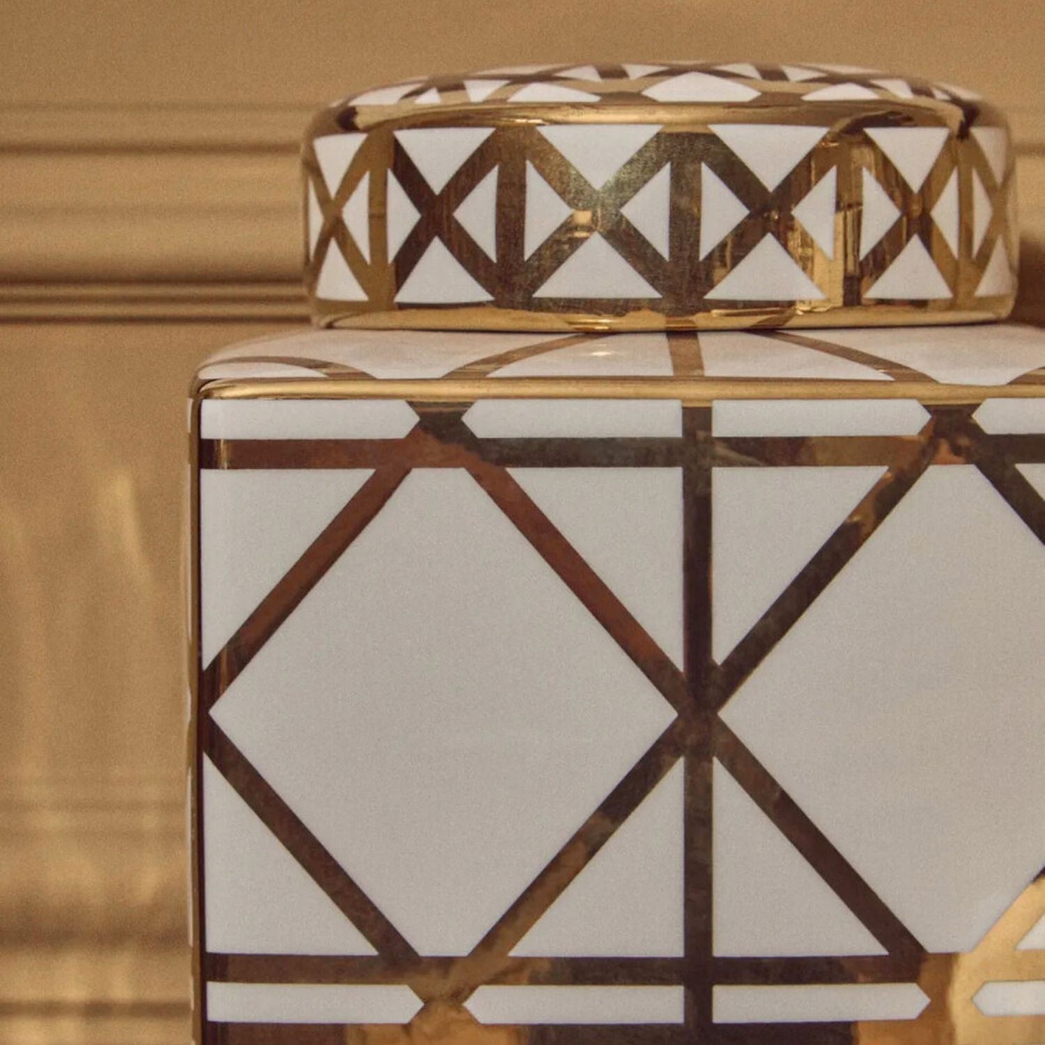 A close-up of a decorative ceramic jar with a white body and lid, featuring a metallic gold geometric lattice pattern.
