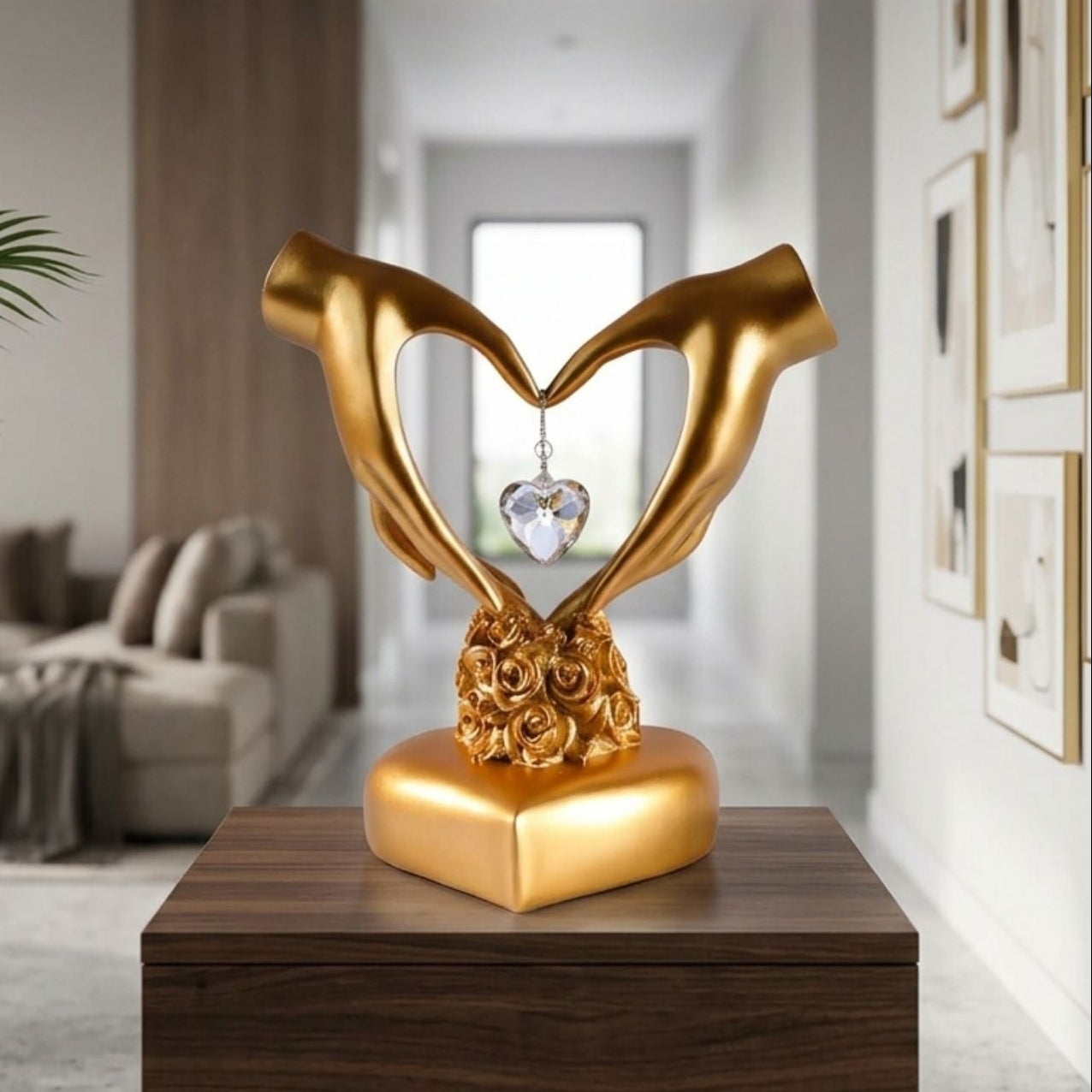A gold-toned decorative sculpture of two hands forming a heart shape above a rose base, with a hanging clear heart crystal, displayed on a dark wooden table in a modern living room setting.