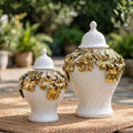 Two white ceramic lidded vases with embossed geometric patterns and raised gold floral accents, displayed outdoors on a woven mat with a blurred garden background.