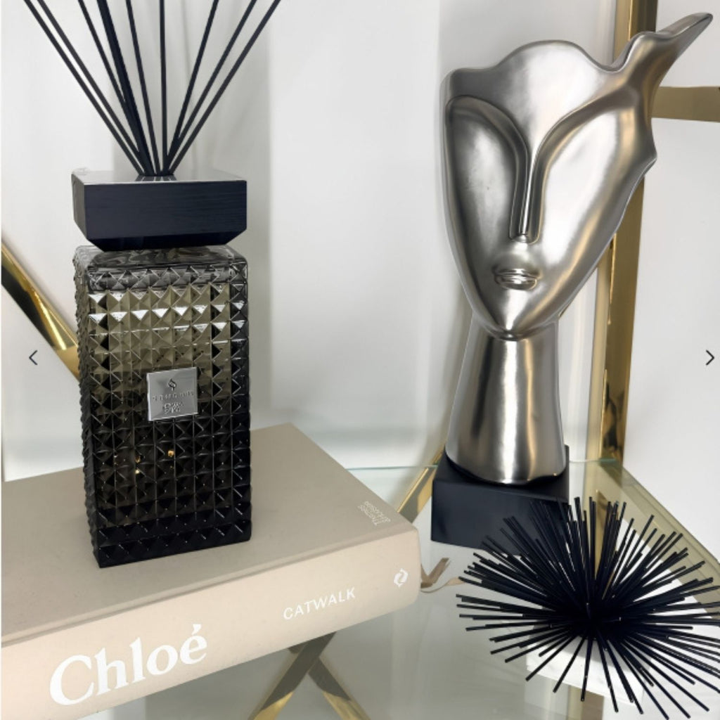 A dark smoky amber glass reed diffuser with a faceted geometric pattern and black reeds, styled on a beige book on a glass shelf next to a silver metal face sculpture.