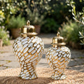 Two decorative ceramic vases with gold lids and a gold-and-white mosaic pattern, displayed outdoors on a stone surface with a blurred garden background.