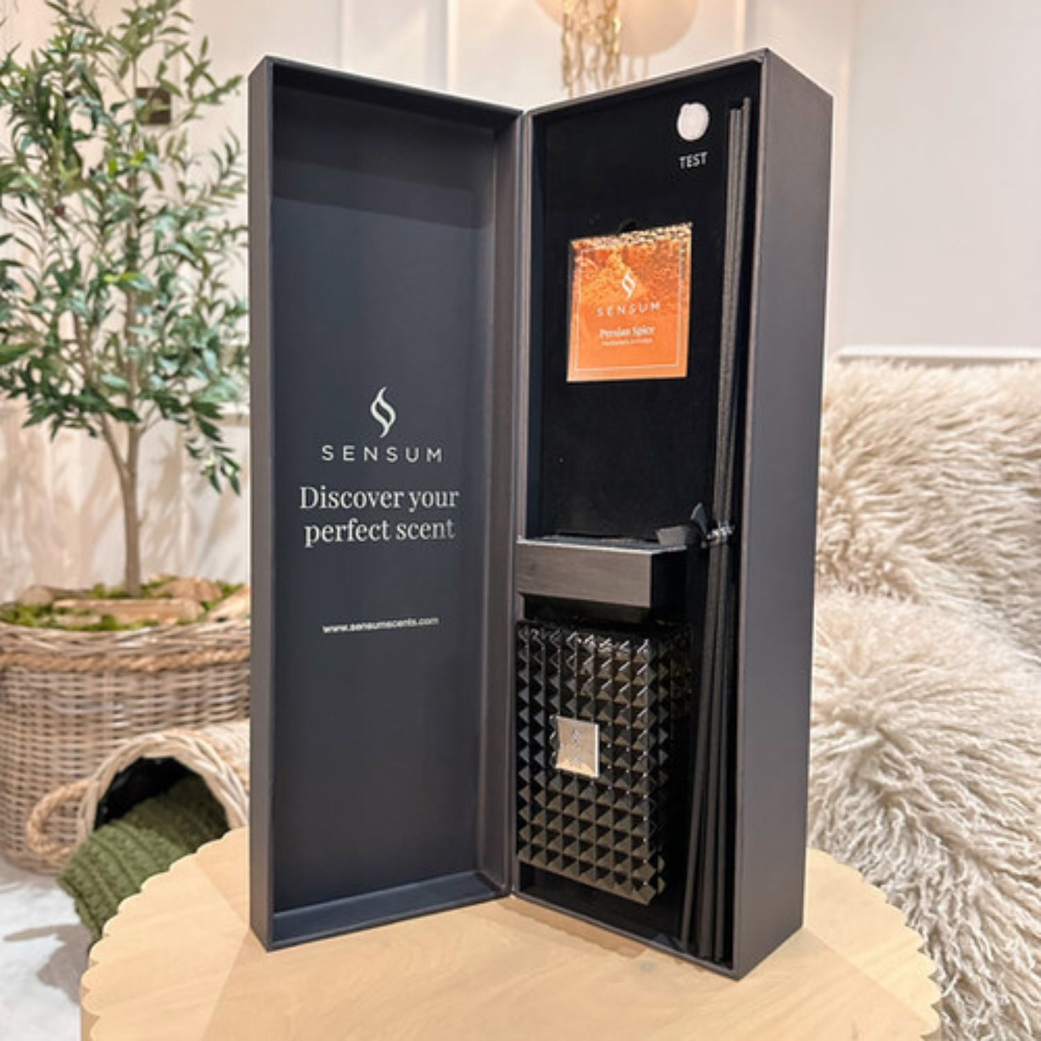 An open SENSUM fragrance gift box displayed on a round wooden table in a softly styled indoor setting. The dark charcoal box is open to reveal a black interior with a textured geometric diffuser or insert and an orange label card.