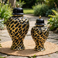 Two decorative ceramic canisters with a glossy black and gold spotted pattern, displayed on a woven mat in an outdoor garden setting.