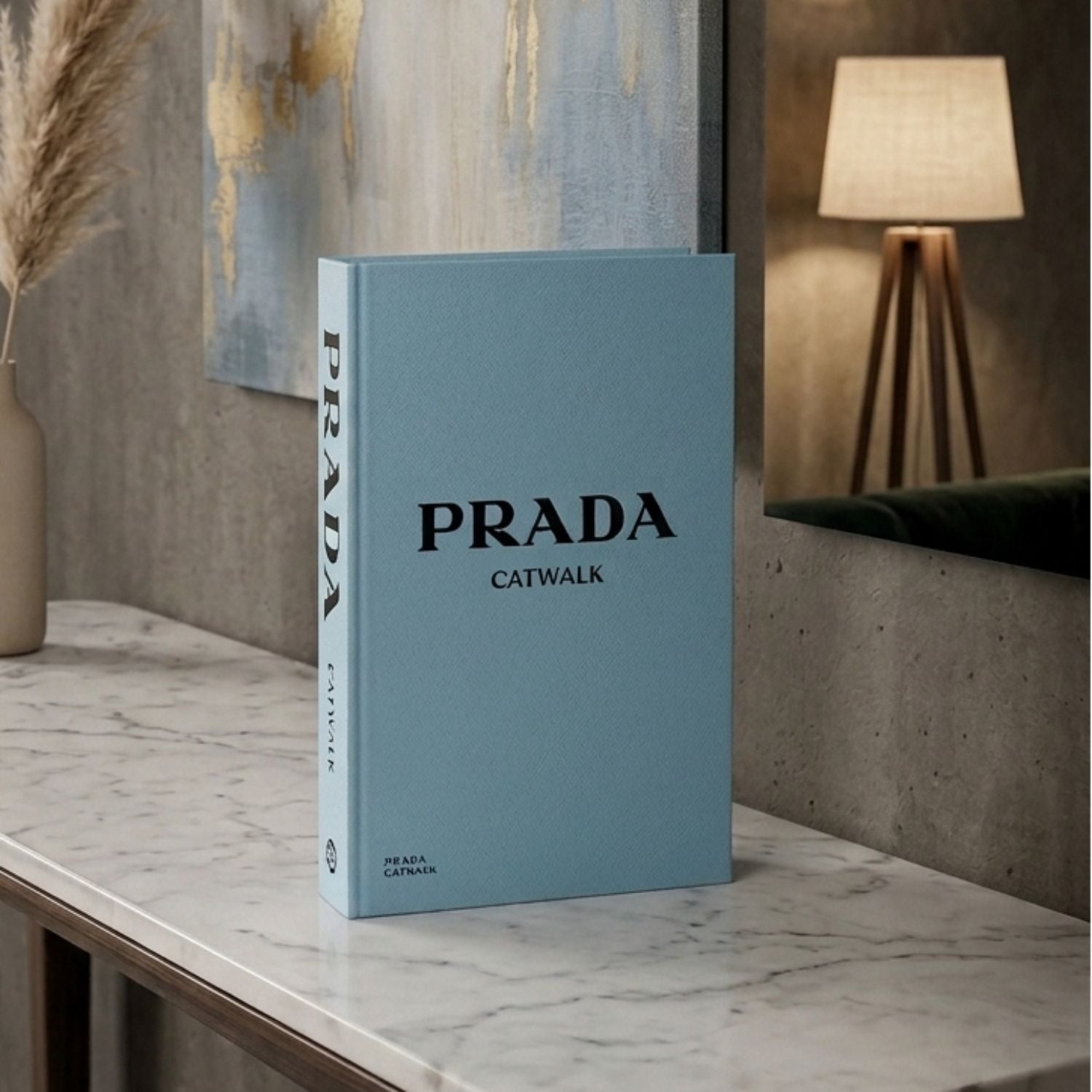 A light blue hardcover book titled 'PRADA CATWALK' standing upright on a marble countertop in a modern interior setting with a lamp and abstract wall art in the background.