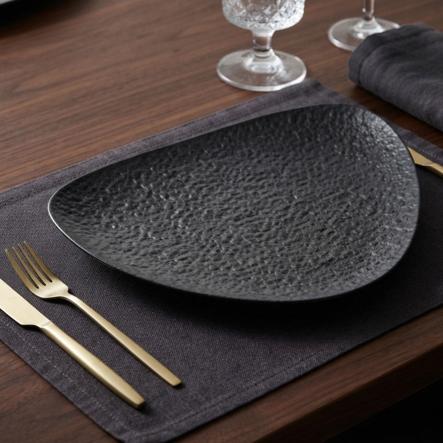 A dark charcoal textured oval serving platter on a dark gray placemat with gold-toned cutlery, set on a wooden table.