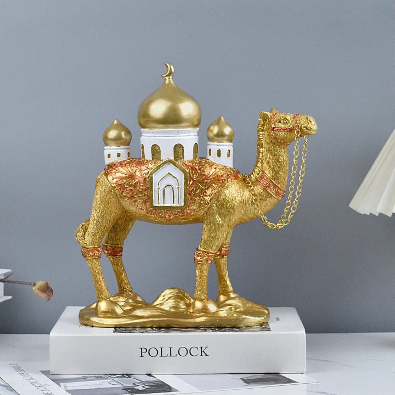 A decorative camel figurine in metallic gold with red and white embroidered details, standing on a white pedestal labeled POLLOCK against a soft gray background.