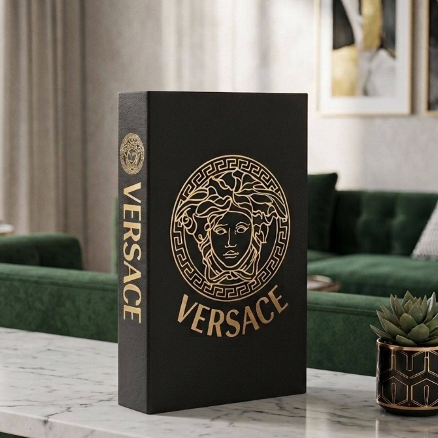 A black Versace decorative book or box standing upright on a marble table in a modern living room setting, featuring a large gold Medusa emblem and the word VERSACE on the front, with the brand name vertically printed on the spine.