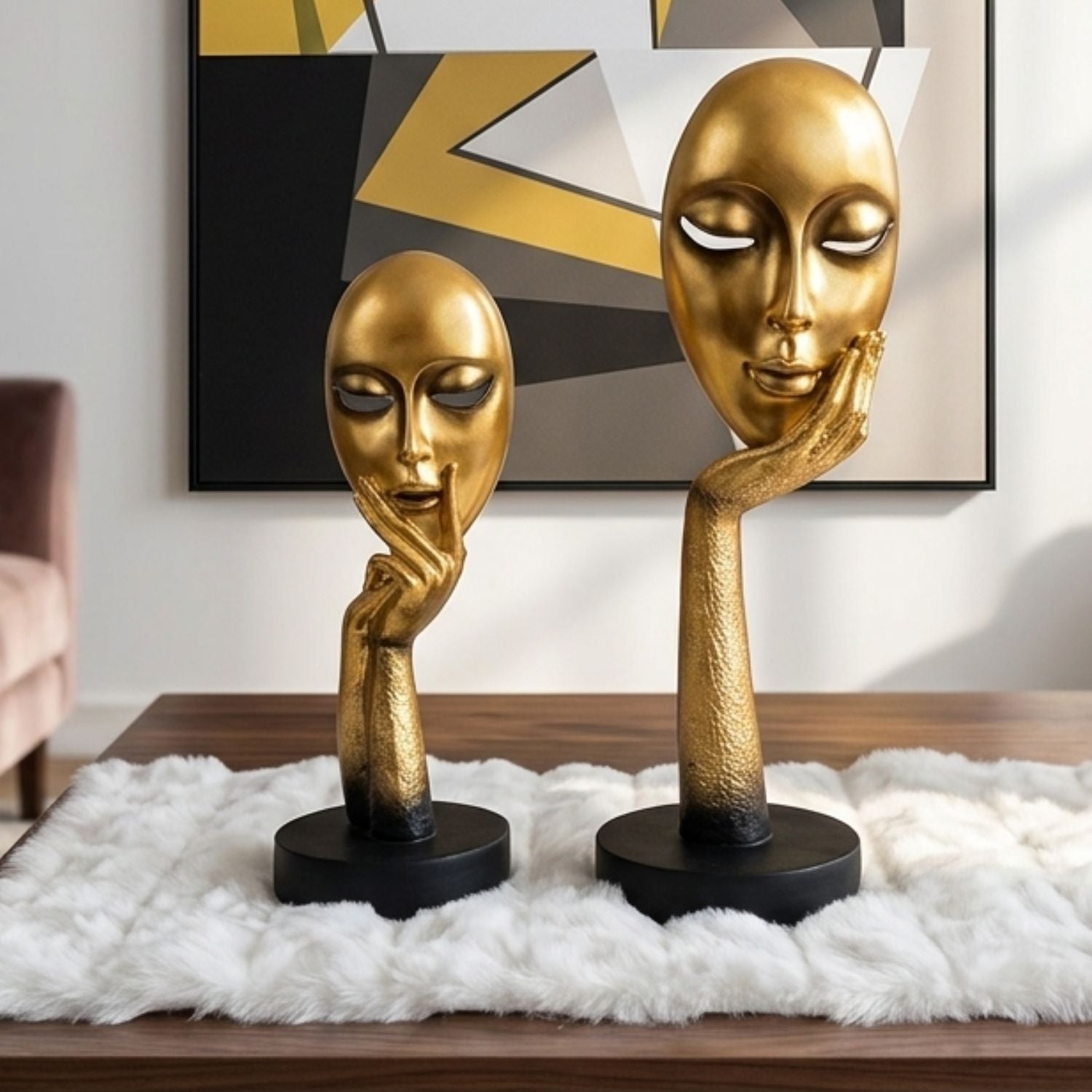 Two gold-toned decorative head sculptures with stylized faces and raised hands, displayed on black bases on a white faux-fur rug in a modern interior setting.