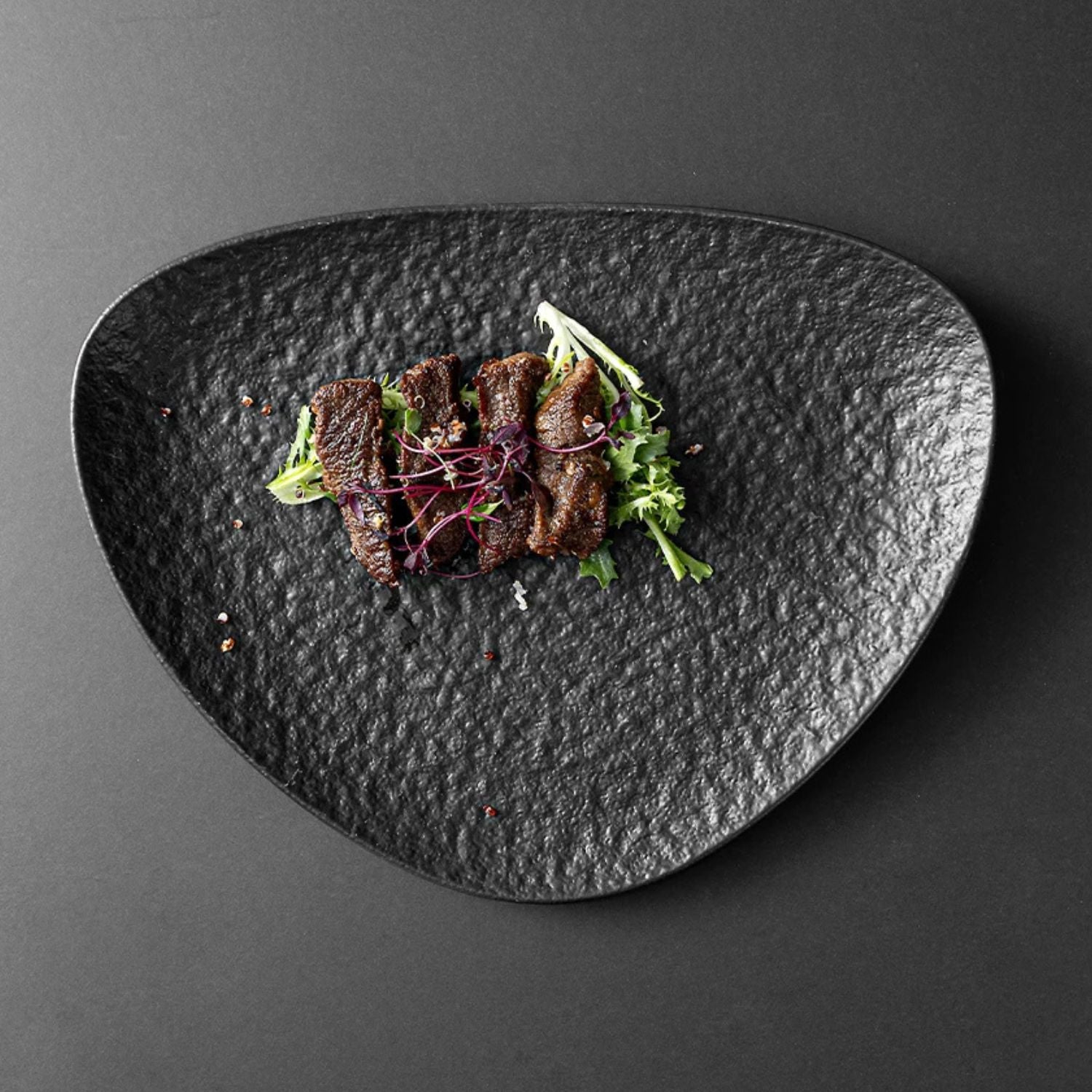 A top-down view of a gourmet food presentation featuring sliced grilled meat on a textured black plate with green and purple garnishes, set against a dark gray background.