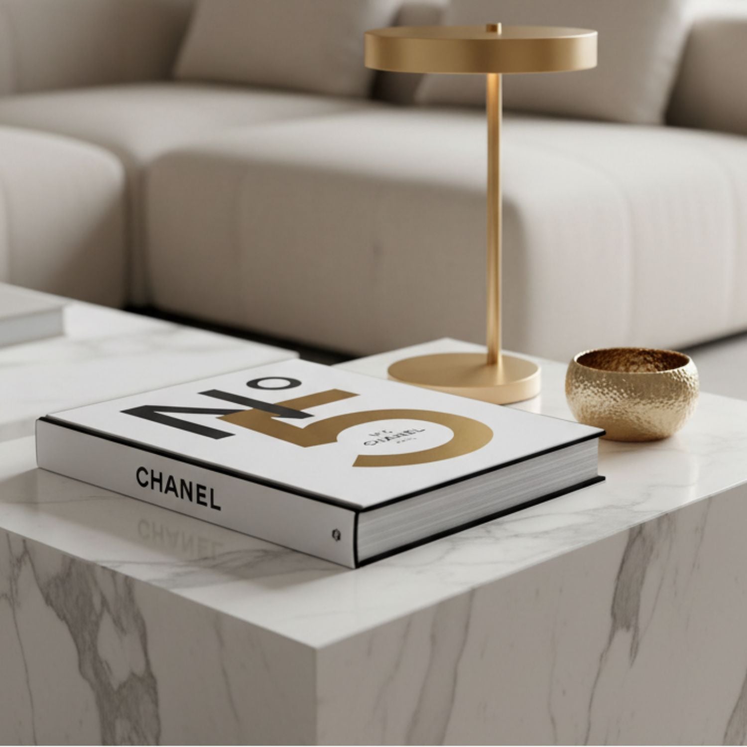 A luxury lifestyle scene featuring a gold modern table lamp and a small gold bowl on a white marble coffee table next to a Chanel No. 5 book, with a beige sectional sofa in the background.