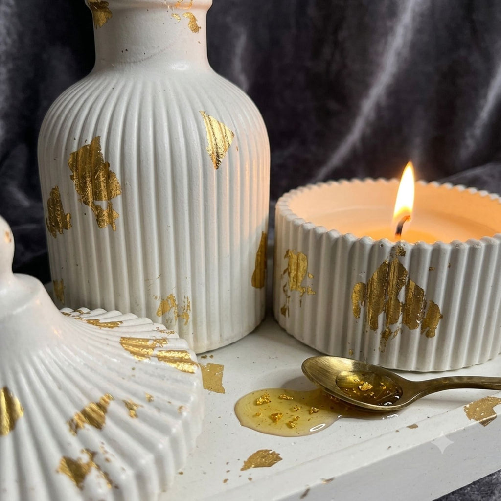 Decorative white ribbed ceramic vessels with scattered gold leaf accents, including a tall vase, a candle holder with a lit candle, and part of a shell-shaped object, arranged on a white surface with a gold spoon and wax pool against a dark draped background.