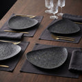 Four dark charcoal textured ceramic plates arranged on dark gray linen placemats on a wooden table, each set with gold-toned cutlery and glassware.