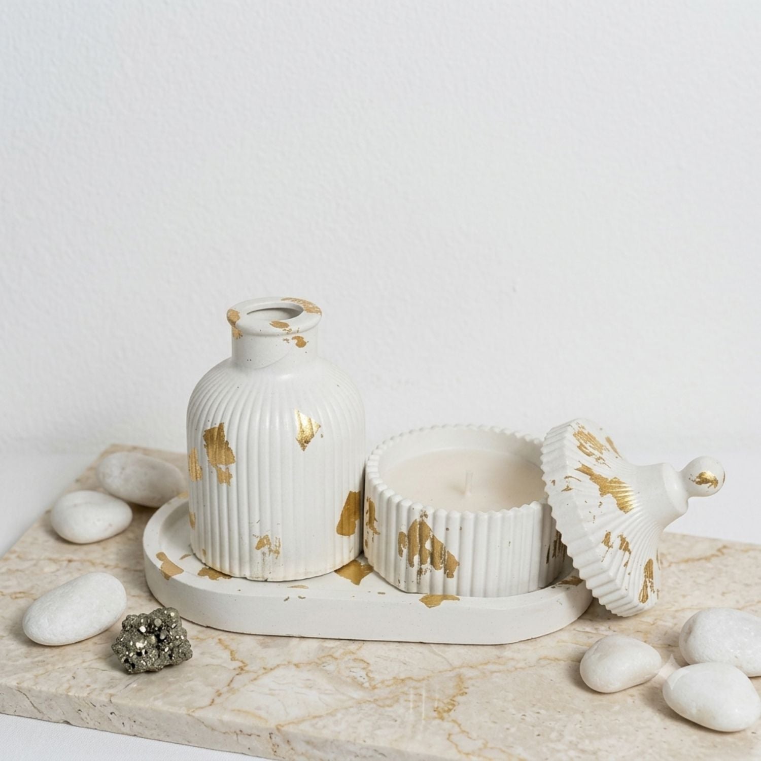 A white ceramic candle set with gold leaf accents, including a ribbed vase-shaped candle holder, a ribbed candle tin, and a matching lid, arranged on a marble tray with decorative white stones against a white wall.