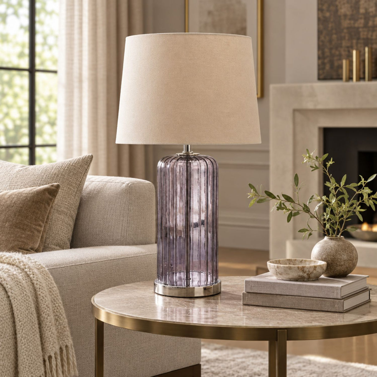 A table lamp with a ribbed lavender-purple glass base and a beige fabric drum shade, styled on a round marble-top side table in a cozy living room setting.