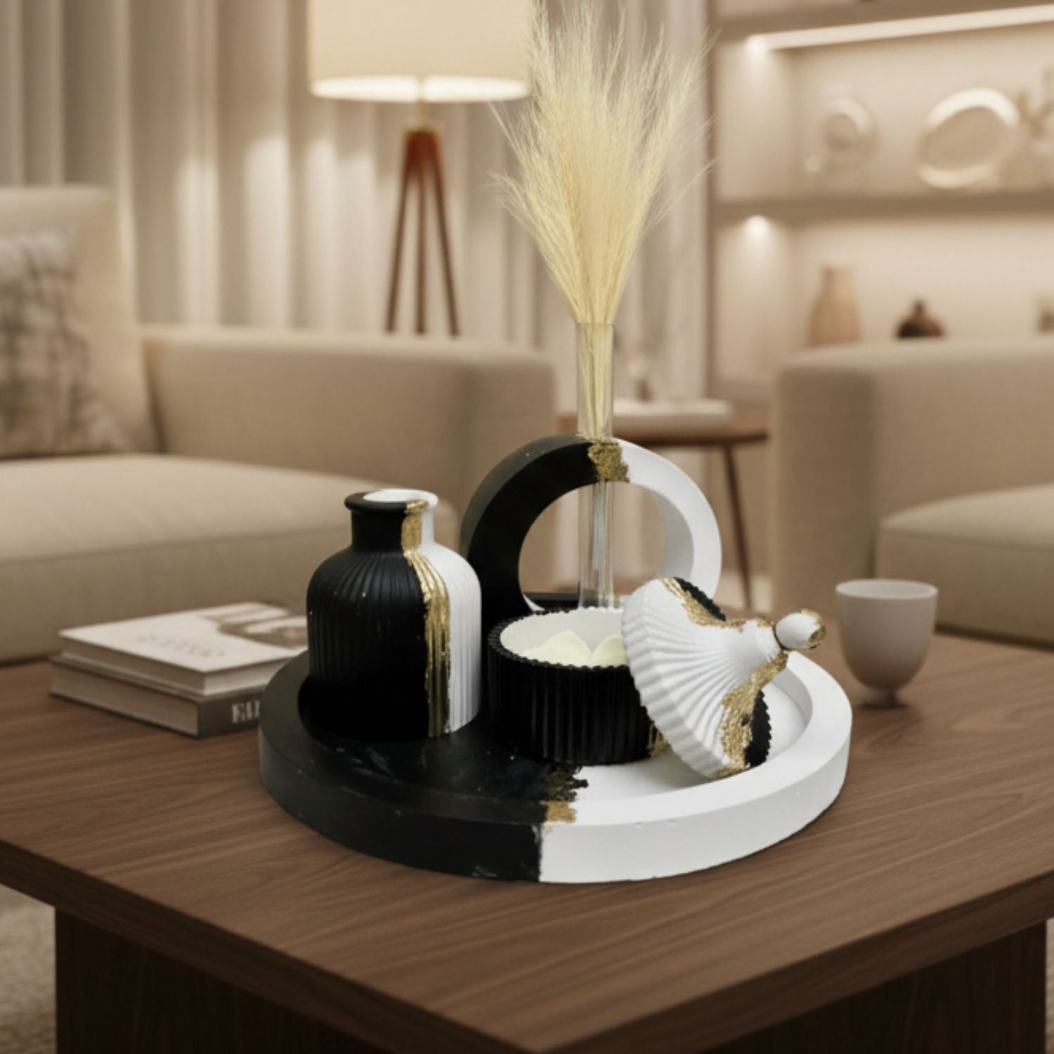Modern black and white decorative tray set on a wooden coffee table in a warm, contemporary living room. The set includes a round white tray with black accents, a ribbed black vase with gold trim, a white shell-shaped decor piece, and a dried grass stem in a black and white arch holder.