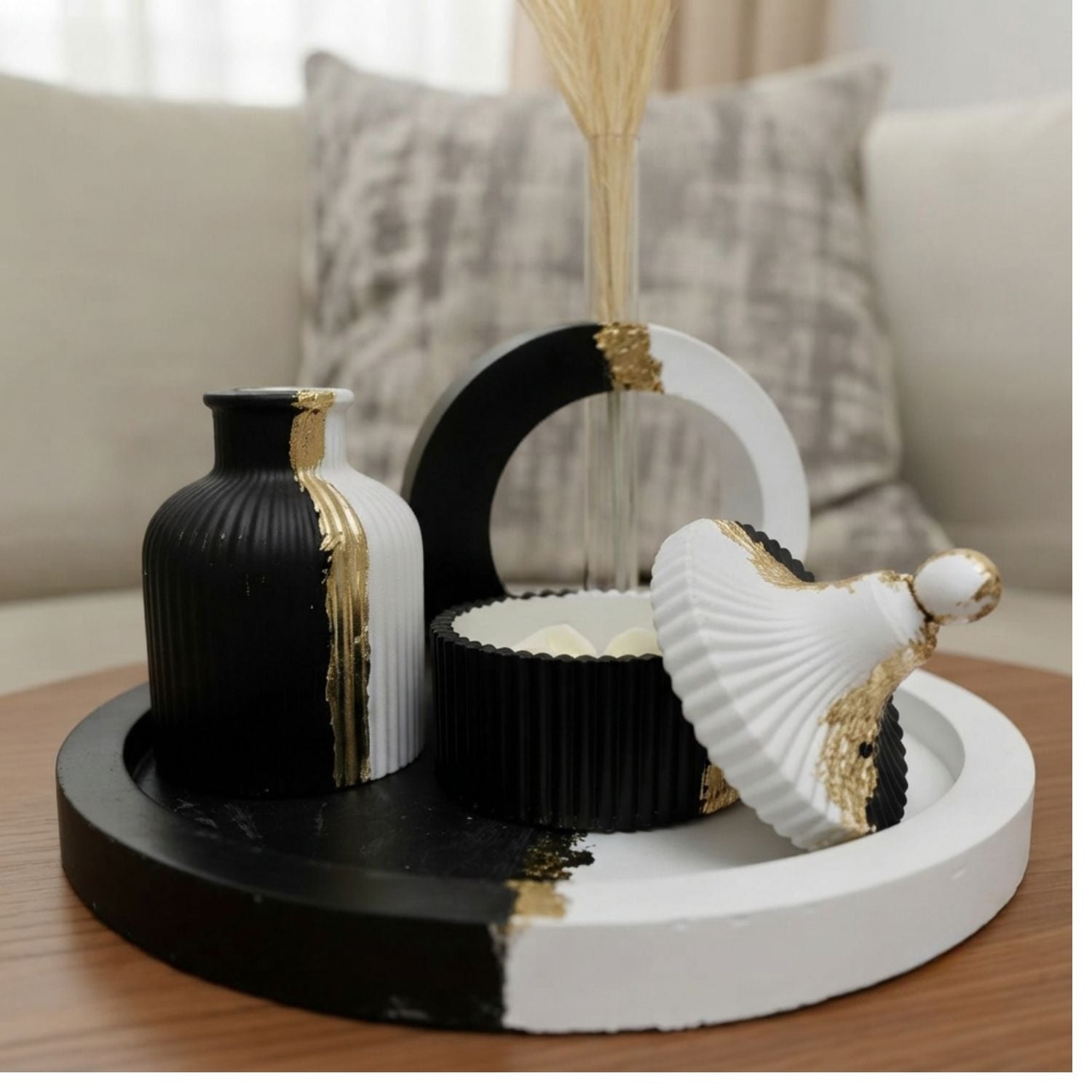 A decorative black and white tray set on a wooden table in front of a sofa, featuring a ribbed black vase with gold leaf accents, a white and black circular sculpture, a white and black ribbed bowl, and a white shell-shaped ornament with gold accents.