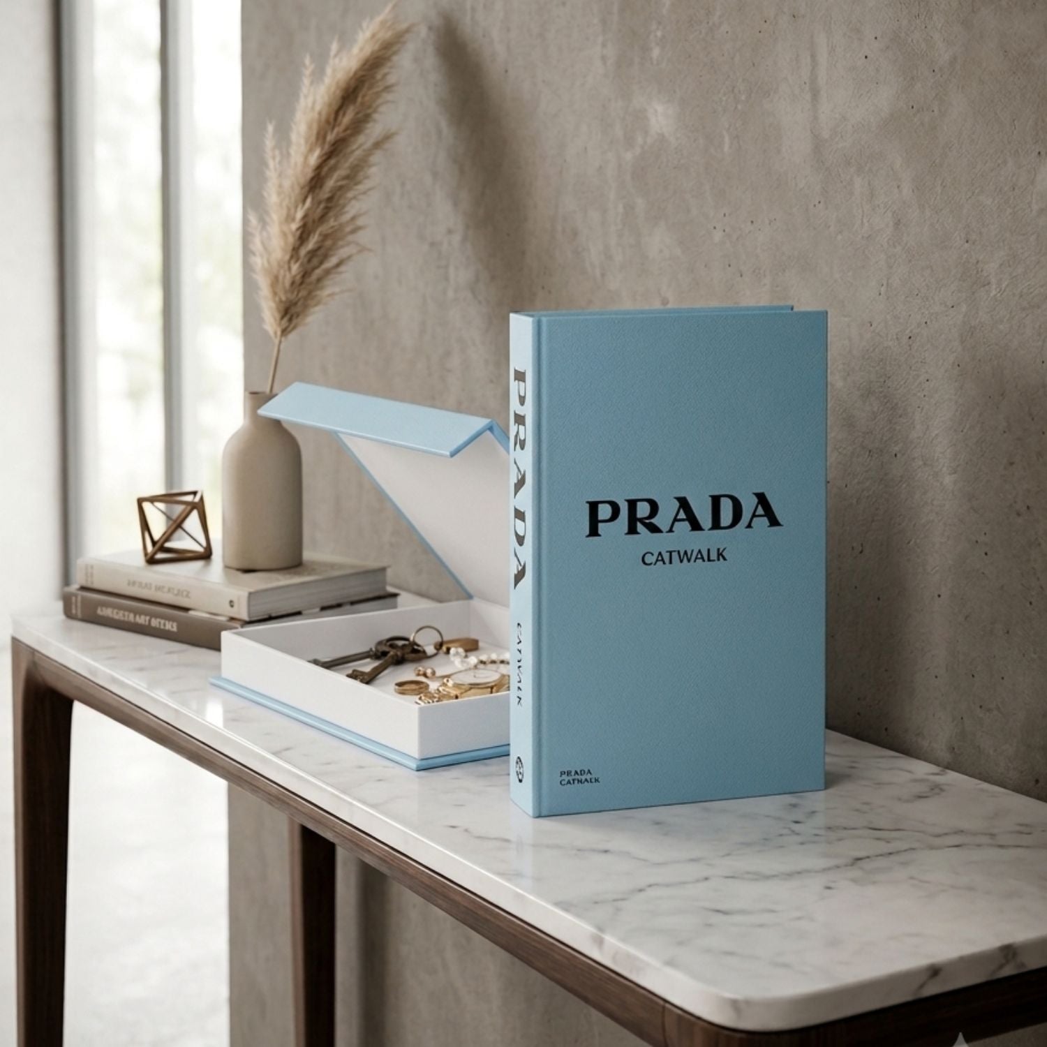 A light blue hardcover book titled 'PRADA CATWALK' stands open on a marble-topped table beside a matching jewelry box, with stacked books, a vase, and dried grass in a softly lit interior.